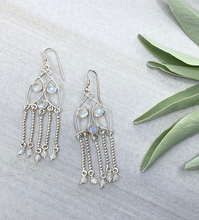 Load image into Gallery viewer, Silver chandelier earrings with moonstone beads on a light surface with green leaves.